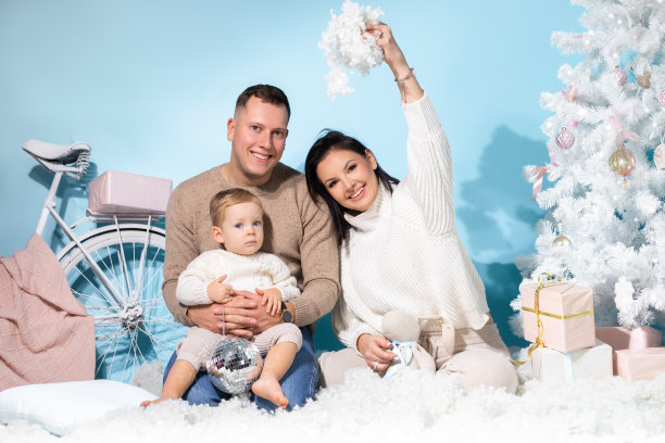 一家三口坐在圣诞照片区，蓝色背景，白色装饰。快乐的妈妈，爸爸和小儿子玩得开心，扔人造雪。新年，节日，礼物图片下载