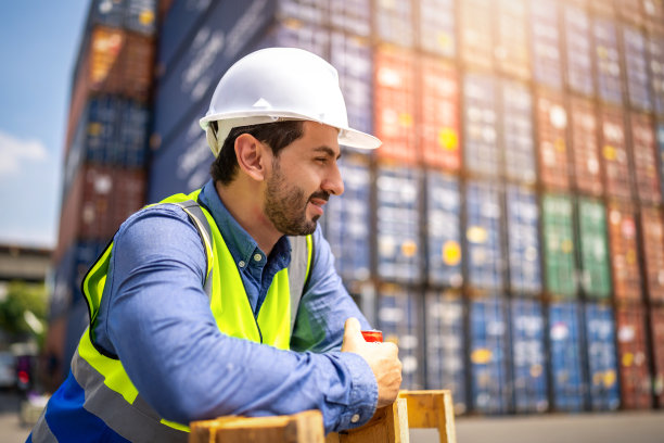 英俊的工程师或工头戴着个人防护装备在日落时检查集装箱存储与货物集装箱背景。物流全球进出口航运的产业概念。图片下载