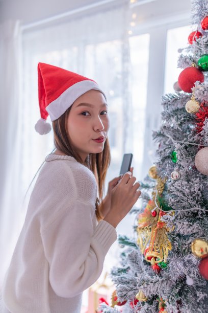 肖像照片的年轻美丽友好的亚洲女性与圣诞老人帽子使用她的手机拍摄的一个漂亮的装饰圣诞树在她的房间图片下载