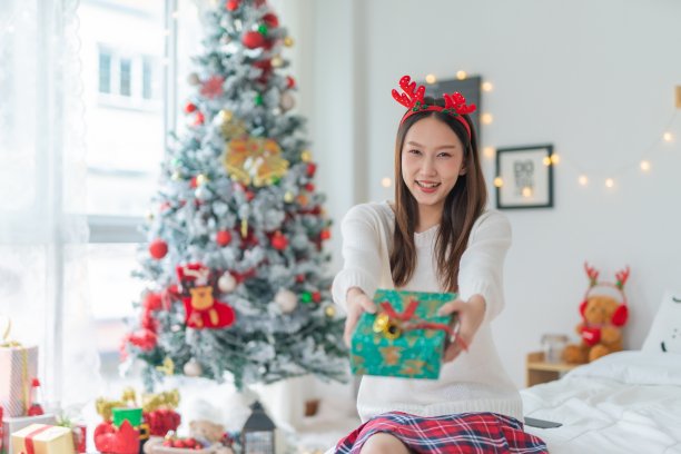 肖像照片的年轻美丽友好的亚洲女性与可爱的圣诞发带愉快地分发礼盒与一个漂亮的装饰圣诞树在背景图片下载
