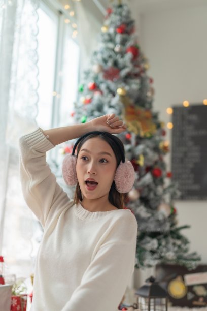 肖像照片的年轻美丽友好的亚洲女性与冬季耳朵罩摆姿势愉快地在她的房间一个漂亮的装饰圣诞树前图片下载