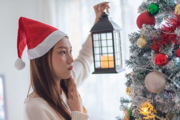 肖像照片的年轻美丽友好的亚洲女性与圣诞老人帽子手持户外灯笼烛光在一个漂亮的装饰圣诞树前图片下载