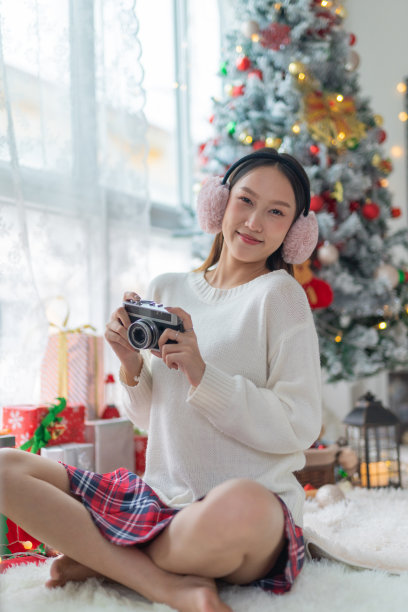 肖像照片的年轻美丽友好的亚洲女性冬季耳罩兴高采烈地抓拍与一个老式胶卷相机在她的房间一个漂亮的装饰圣诞树前图片下载