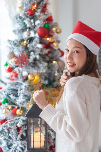 肖像照片的年轻美丽友好的亚洲女性与圣诞老人帽子手持户外灯笼烛光在一个漂亮的装饰圣诞树前图片下载