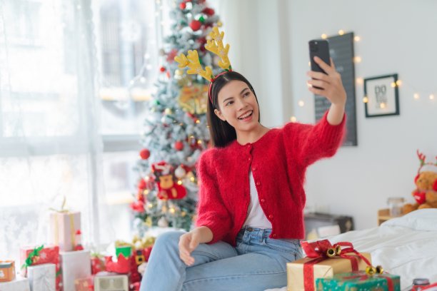 肖像照片的年轻美丽友好的亚洲女性与可爱的圣诞发带愉快地自拍与她的智能手机在她的房间一个漂亮的装饰圣诞树前图片下载