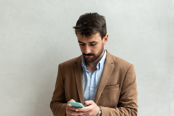 年轻的企业家站在室内靠墙的地方用智能手机收发短信。一本正经的商人给他的同事们发送关于统计数据和计划的电子邮件。图片下载