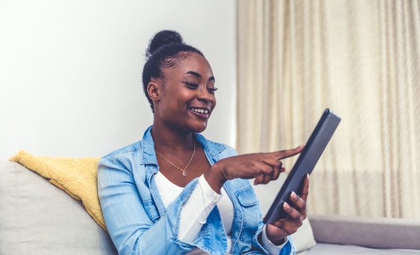 年轻的非洲裔美国妇女坐在家里的沙发上微笑，挥手问候，说话，在pad上视频通话。快乐的混血女性在平板电脑上有网络摄像头数字虚拟事件。通信的概念。图片下载
