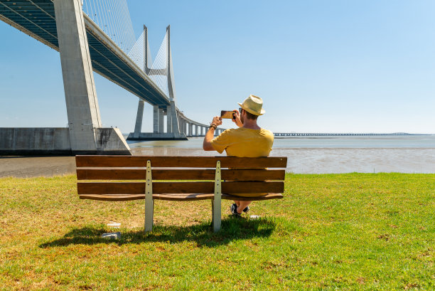 一名年轻男子坐在长凳上，在葡萄牙里斯本的瓦斯科达伽马桥上拍照。图片下载