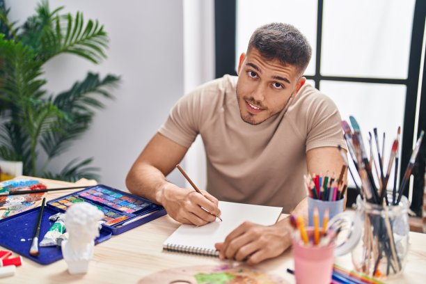 年轻的西班牙男子艺术家微笑自信地在艺术工作室的笔记本上画画图片下载