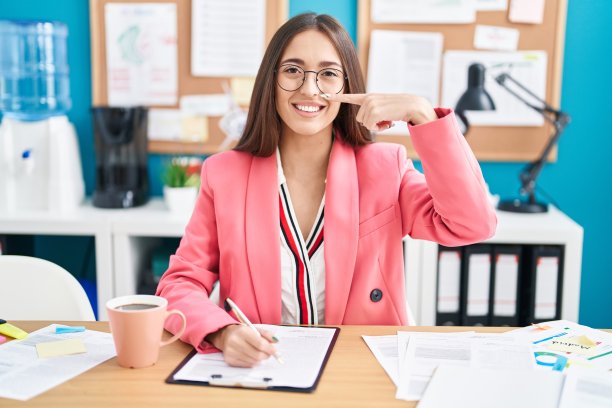 年轻的西班牙女性在办公室工作，戴着眼镜，用手和手指指着脸和鼻子，微笑愉快。美丽的概念图片下载