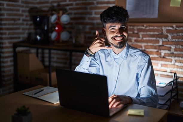 年轻的西班牙人，留着胡子，晚上在办公室工作，微笑着用手和手指做电话手势，就像在讲电话。沟通的概念。图片下载