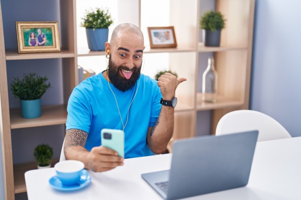 年轻的西班牙男子，留着胡子和纹身，用智能手机做视频电话，拇指朝上，张开嘴笑得很开心图片下载