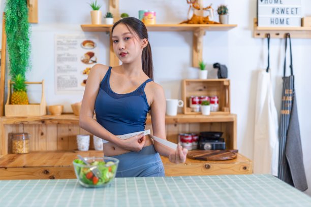 照片的年轻美丽的亚洲妇女女士穿着运动服快乐做腰测量检查她的训练，锻炼和饮食的结果。她家厨房桌上的樱桃番茄沙拉是为了减肥和健康的生活方式图片下载
