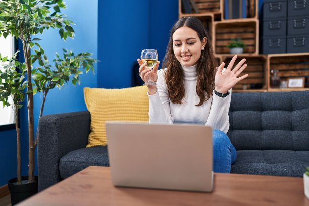 年轻的西班牙妇女做视频电话喝白葡萄酒，看起来积极和快乐的站着，微笑与自信的微笑露出牙齿图片下载