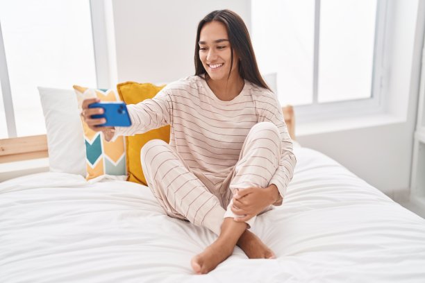 年轻美丽的西班牙女子坐在卧室的床上用智能手机自拍图片下载