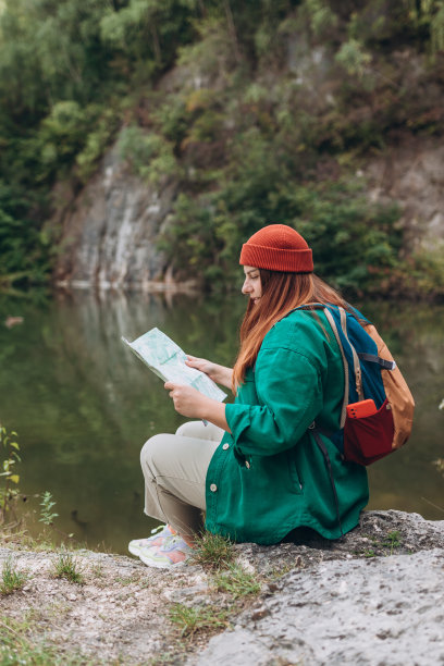 时髦的潮女人拿着纸地图，背着双肩包，戴着红帽子，在自然湖的背景下放松。旅行和漫游的概念。令人惊叹的寒意时刻。全身照片图片下载