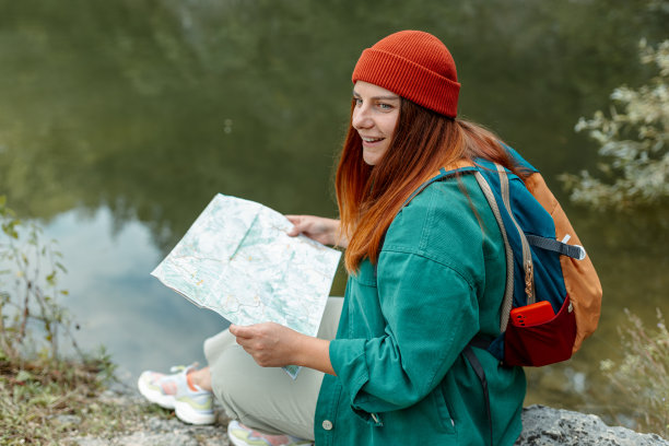 时髦的潮女人拿着纸地图，背着双肩包，戴着红帽子，在自然湖的背景下放松。旅行和漫游的概念。令人惊叹的寒意时刻。全身照片图片下载