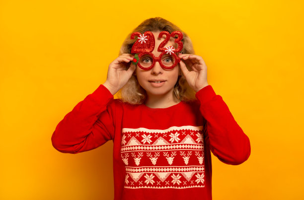 惊讶的年轻金发女子拿着派对眼镜，穿着装饰图案的红色圣诞毛衣，看着黄色背景上的相机。

2023年新年快乐概念。图片下载