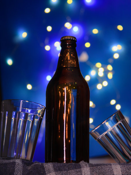 这是一张肖像式的照片，一瓶啤酒和两个玻璃杯放在毛巾上，蓝色背景上的散景灯，参考了圣诞节和新年的聚会。图片下载