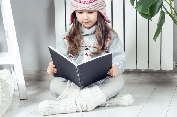忧郁严肃可爱的小女孩，戴着羊毛帽子，穿着暖和的衣服，雪人在取暖散热器旁边看书或记事本图片下载