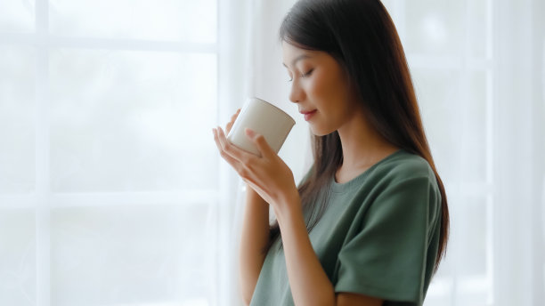 年轻的亚洲妇女站在窗边，拿着杯子在家里的卧室，她喝牛奶后，早晨醒来图片下载