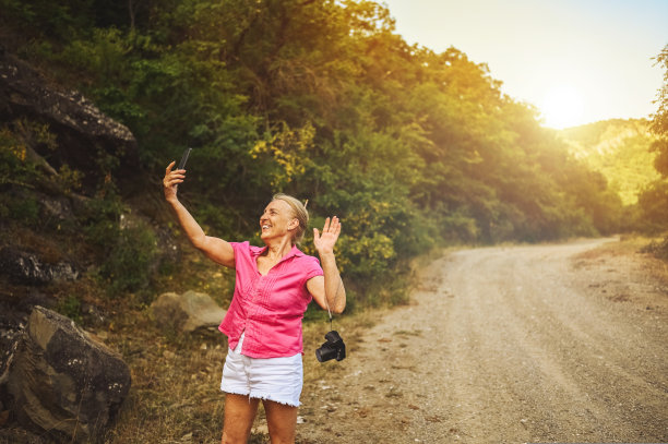 兴奋快乐的资深女性背包客走在夏季森林路户外在日落时分。老苗条的女士旅行的照片相机。积极的退休假期理念。温暖的过滤器。图片下载