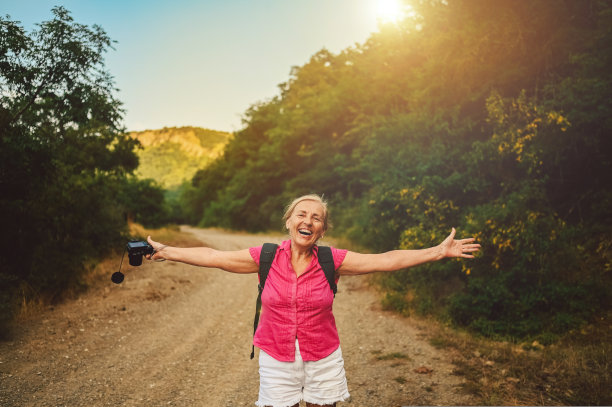 兴奋快乐的资深女性背包客走在夏季森林路户外在日落时分。老苗条的女士旅行的照片相机。积极的退休假期理念。温暖的过滤器。图片下载