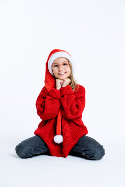 一个年轻漂亮的女孩戴着圣诞帽坐在白色的地板上，微笑着看着镜头，双手托着下巴。孤立的背景图片下载