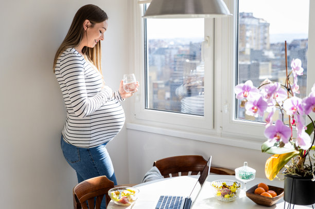 孕妇站在家里的窗前。端着一杯水。图片下载