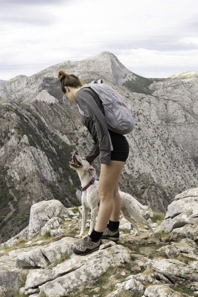 垂直镜头中，一个年轻的白人女孩穿着山装，背着灰色背包，正在抚摸她的狗。背景是邻近的一座美丽的山。徒步旅行和登山的壁纸。图片下载