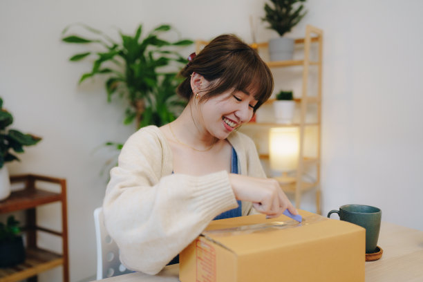年轻的亚洲妇女从纸板箱中打开新购买的衣服，这是从她的网上购物零售送货在家。图片下载