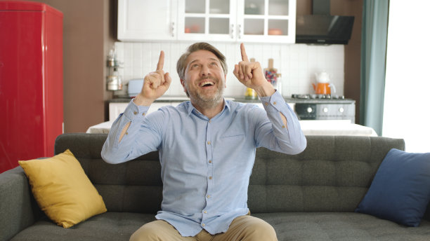 年轻男子坐在公寓里舒适的沙发上，手指指向屏幕上方的摄像机。图片下载