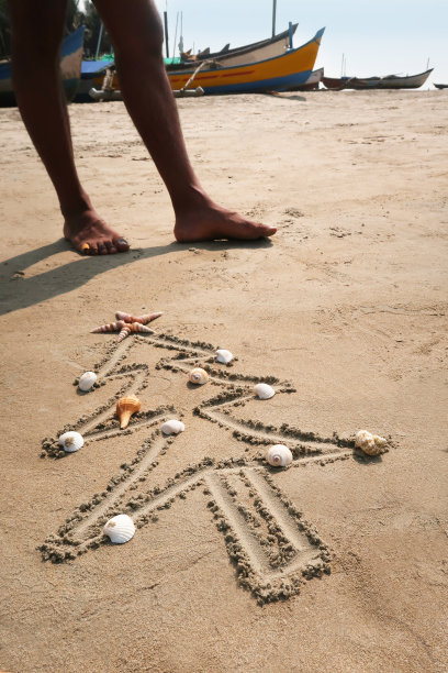 图片中，一个无法辨认的人站在阳光明媚的海滩上，用贝壳装饰的棍子画在沙子上的圣诞树旁，圆锥形贝壳的圣诞星，渔船背景，重点放在前景，南半球的圣诞节概念图片下载