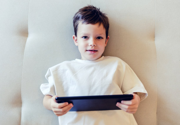 男孩在家躺在床上玩平板电脑游戏。白人小男孩坐在沙发上玩平板电脑游戏。图片下载