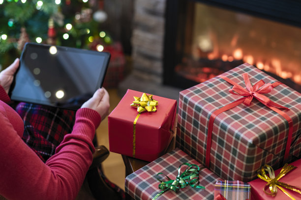 圣诞节。女人在毛衣使用平板电脑搜索礼物的想法坐在桌子旁壁炉和圣诞树。概念图片下载
