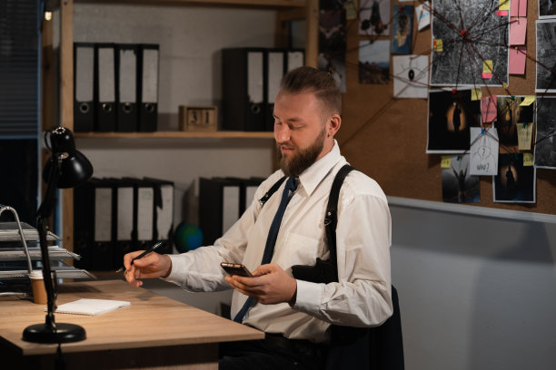 一位职业侦探晚上在办公室调查一起谋杀案。他坐在桌子旁，手里拿着智能手机，在网上阅读新闻。图片下载