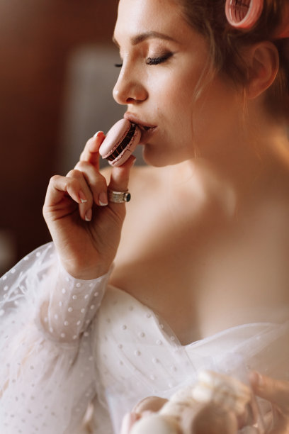 一个美丽的微笑的年轻黑发女人明亮的化妆吃美味的法国马卡龙或马卡龙的美丽肖像。女孩穿着白色的裙子，戴着粉色的卷发，坐在床上放松图片下载