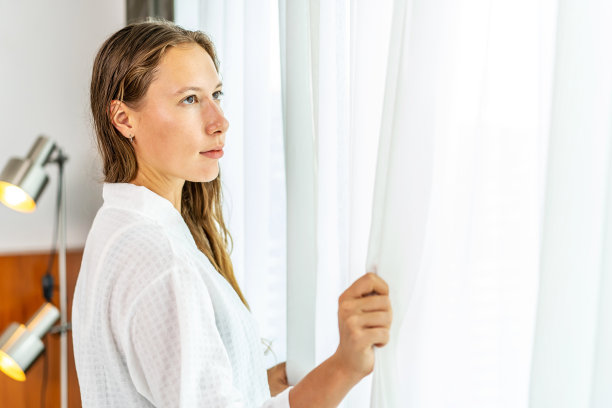 美丽的年轻白人女性在清晨醒来，清新地站在窗前，打开白色的窗帘。图片下载
