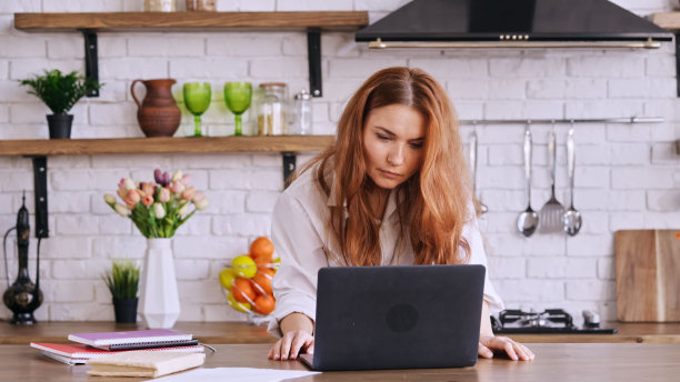 姜头发的女孩站在她的厨房桌子前工作，她的笔记本电脑放在桌子上图片下载