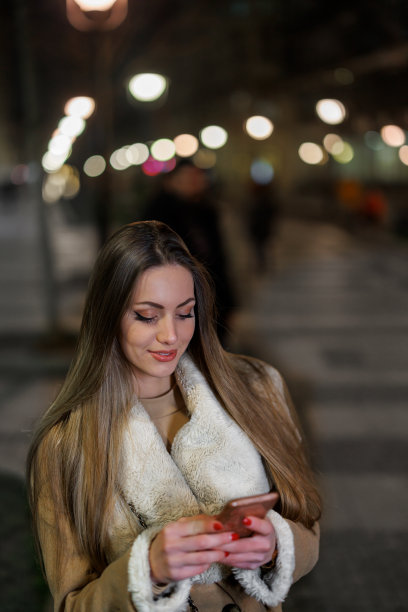 一个年轻的女人晚上走在城区里，用手机发短信。图片下载
