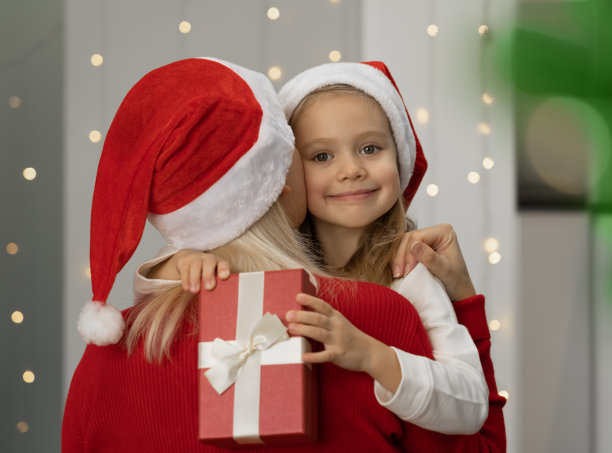 小女孩抱着盒子和妈妈在圣诞树旁微笑。新年礼物图片下载