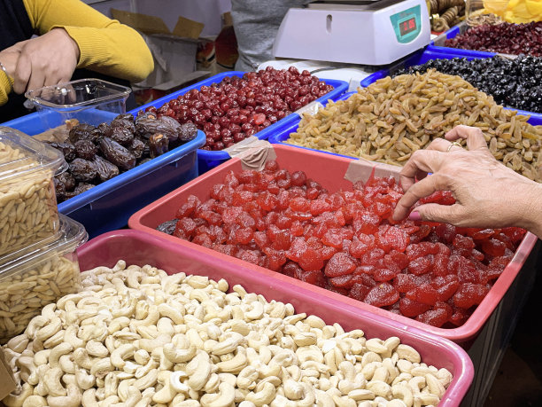 全帧图像无法识别的小贩和顾客在市场摊位上展示，成堆的坚果和干果的塑料容器，准备按重量出售，腰果，蜜饯樱桃，干枣，健康食品零食，高架视图，重点在前景图片下载