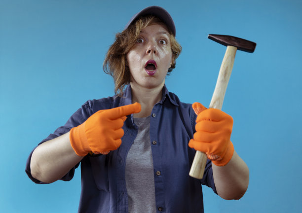 一个穿着衬衫，戴着橙色手套的帽子的女人在蓝色的孤立背景上展示了一把木锤子。女人画像成人。女人努力工作图片下载
