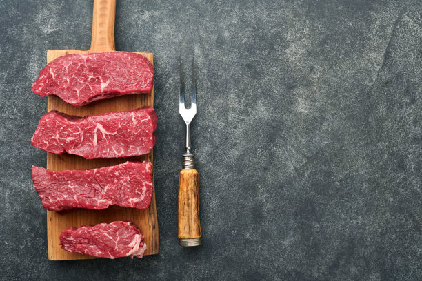 生的牛排。顶部刀片牛排在木头燃烧板上与香料，迷迭香，蔬菜和黑色背景的烹饪材料。前视图。副本的空间。图片下载