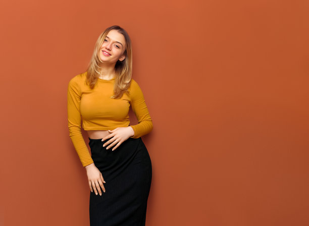 美丽的摆姿势的金发女士大乳房，握着她的手在她的肚子上，心满意足地看着相机与她的头倾斜，在黄色衬衫和黑色长裙橙色坚实的背景与复制空间。图片下载