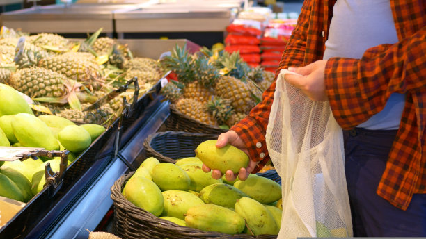 一个人在超市的水果区挑选最美味成熟的芒果，把它们放进一个白色的细绳袋中。遵循适当健康的饮食。男人买水果的概念。图片下载
