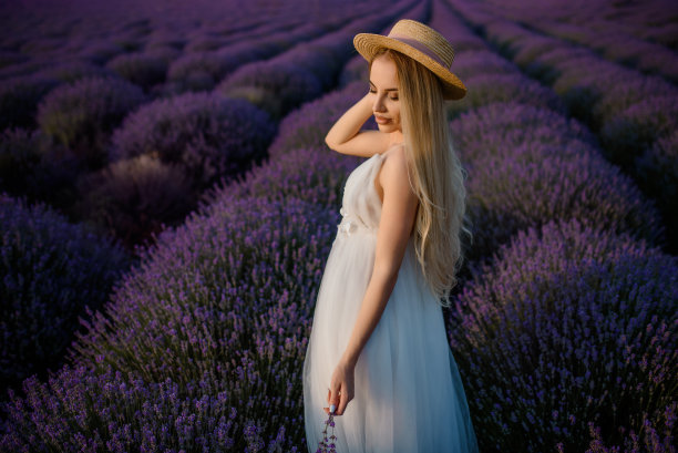 一个年轻美丽的女人，穿着白色的裙子，戴着一顶帽子，走在薰衣草田。图片下载