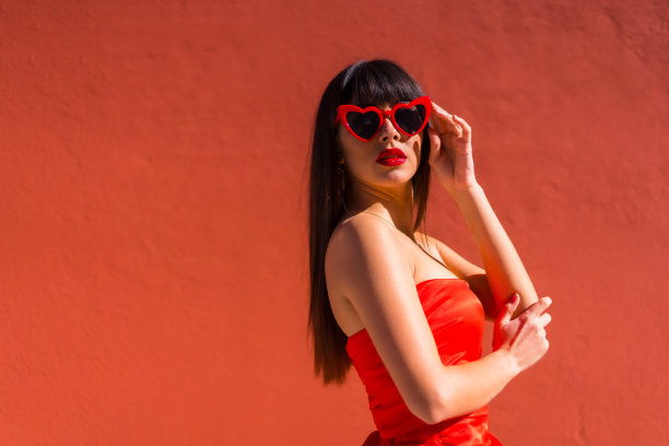 深色头发的女孩在红色背景与红心眼镜和红色紧身连衣裙，情人节微笑图片下载