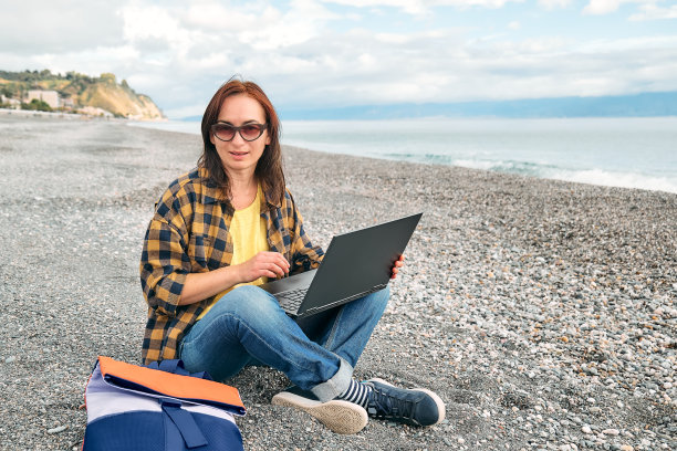穿着温暖的格子衬衫，拿着笔记本电脑的女子坐在海边的沙滩上，在户外工作。现代生活方式、人际关系、博客、商业、自由职业。图片下载