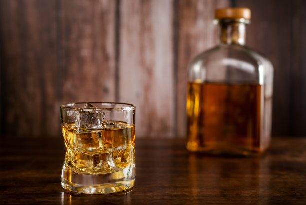 一杯加冰块的威士忌，一瓶威士忌的背景模糊了，背景是木制的。图片下载